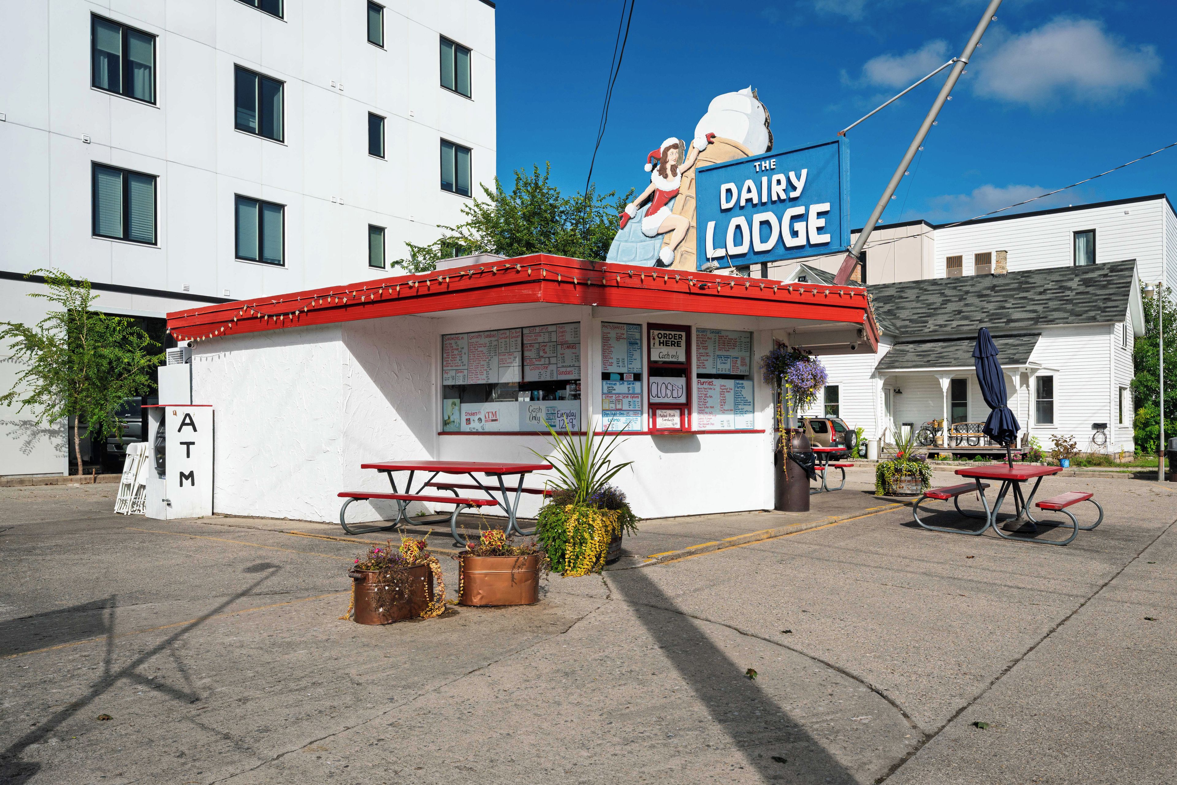 Ein kleines, freistehendes Gebäude mit rotem Dach und weißen Wänden, mit einem Schild für 'The Dairy Lodge'. Draußen stehen Picknicktische und es gibt ein Schild für einen Geldautomaten.