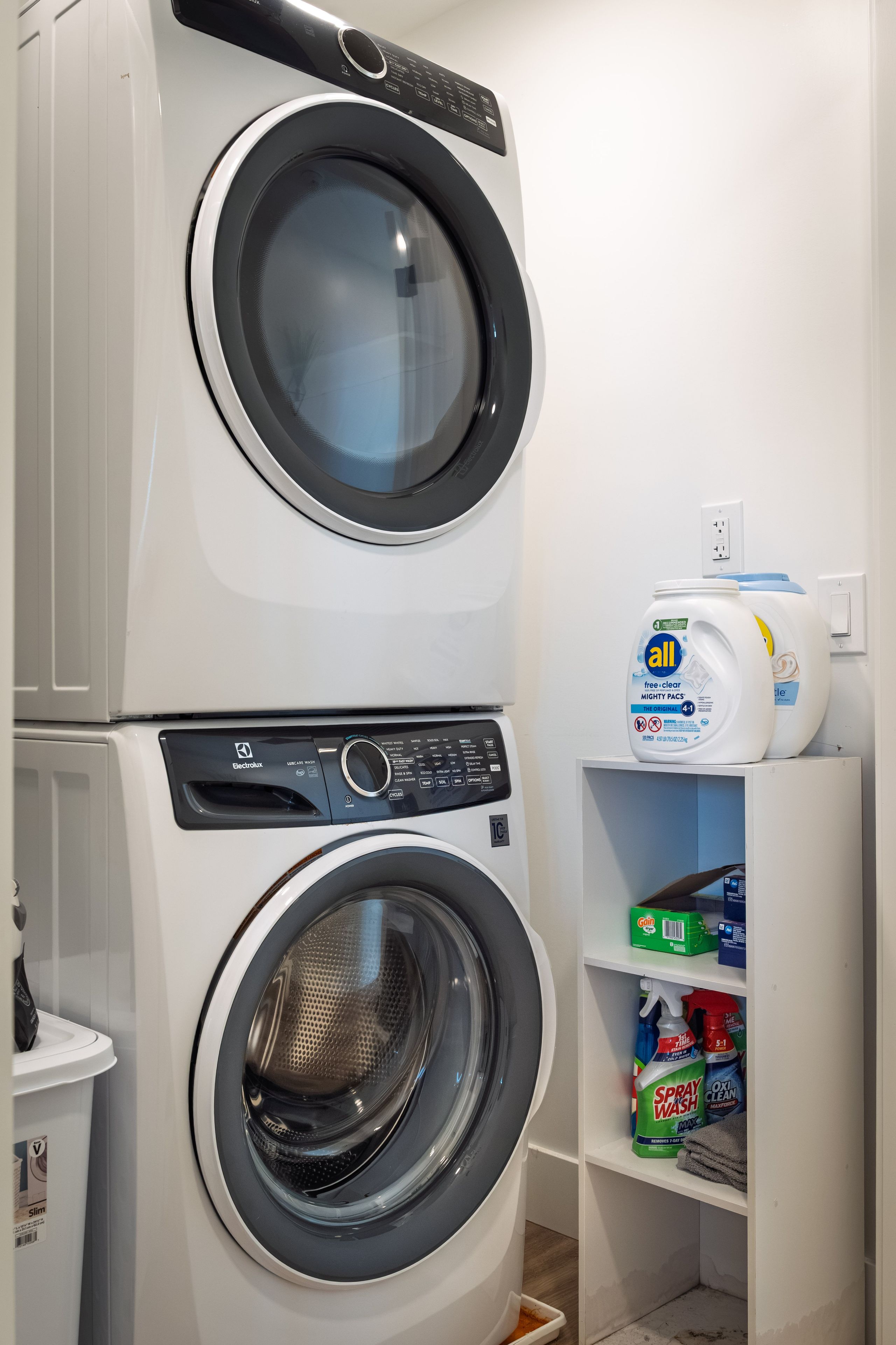 Een gestapelde wasmachine en droger met een plank vol schoonmaakmiddelen.