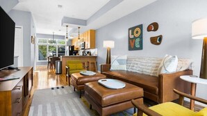 Ein modernes Wohnzimmer mit einer Couch, Poufs und einem Fernseher. Im Hintergrund befindet sich eine Küche mit Holzschränken und einem Essbereich.