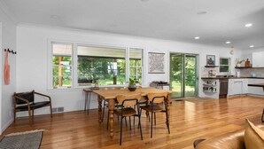 A spacious dining area with a wooden table and chairs, large windows, and a view of greenery outside. - Bayside Bungalow: West Bay Views Minutes to TC (Traverse City)