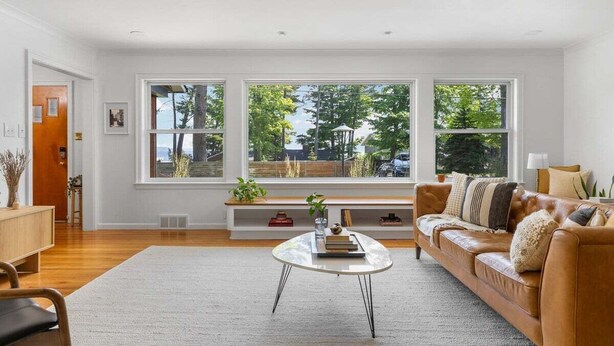 A spacious living room with a large window, a leather sofa, a glass coffee table, and a wooden bench with plants. - Bayside Bungalow: West Bay Views Minutes to TC (Traverse City)