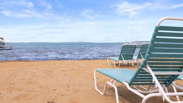 A beach with green lounge chairs and a boat docked in the water. - #01 Beach Bungalow at Lakeshore Resort (Traverse City)