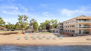 A beachfront hotel with multiple buildings, a sandy beach with sun loungers, and a clear sky. - #01 Beach Bungalow at Lakeshore Resort (Traverse City)