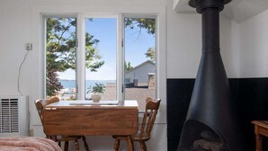 Une pièce avec une table et des chaises en bois, un poêle en bois noir et une fenêtre donnant sur des arbres et un bâtiment.