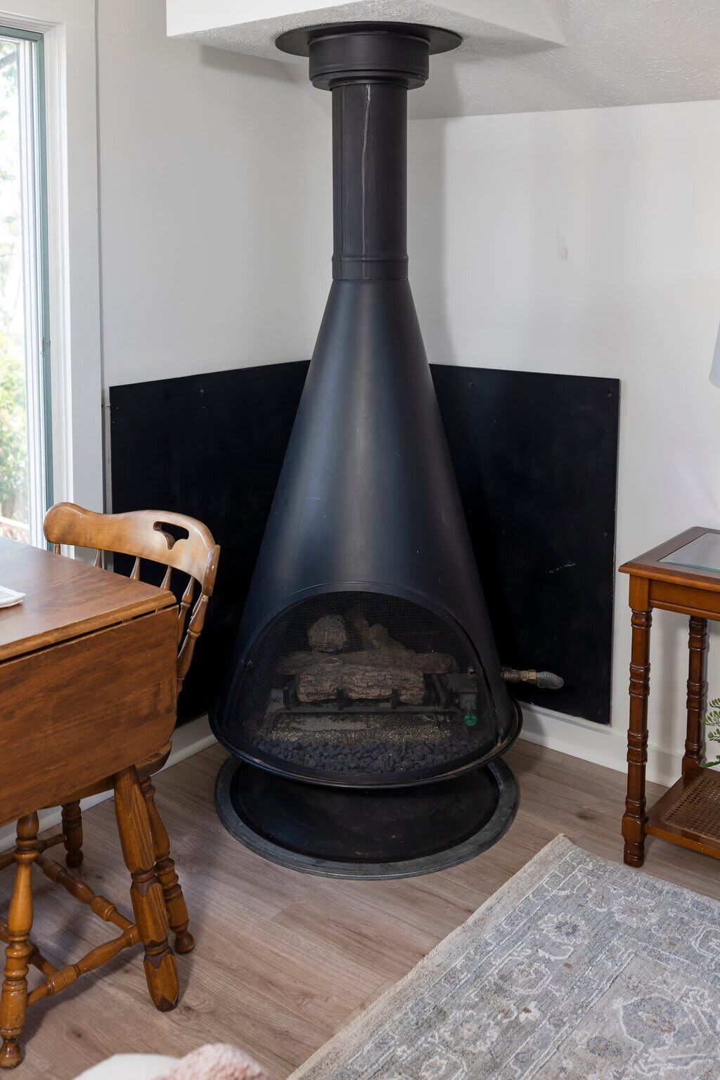 A wood-burning stove with a chimney, a dining table, chairs, and a rug in a room with a window.