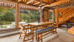 A wooden dining area with a table and benches, large windows, and a view of greenery outside. - Boardman River Cabin (Traverse City)