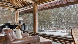 A cozy cabin interior with a leather sofa, a piano, and large windows offering a snowy forest view. - Boardman River Cabin (Traverse City)