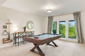 A pool table in a room with a clock on the wall, a plant, and a window with curtains.
