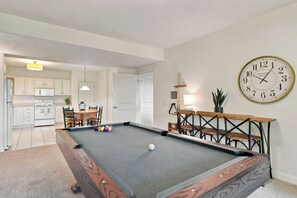 A pool table in a room with a clock on the wall, a dining area, and a kitchen in the background.
