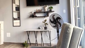 En moderne stue med en grå sofa, et fladskærms-tv monteret på væggen, et lille bord med en ventilator, og et vindue med udsigt.