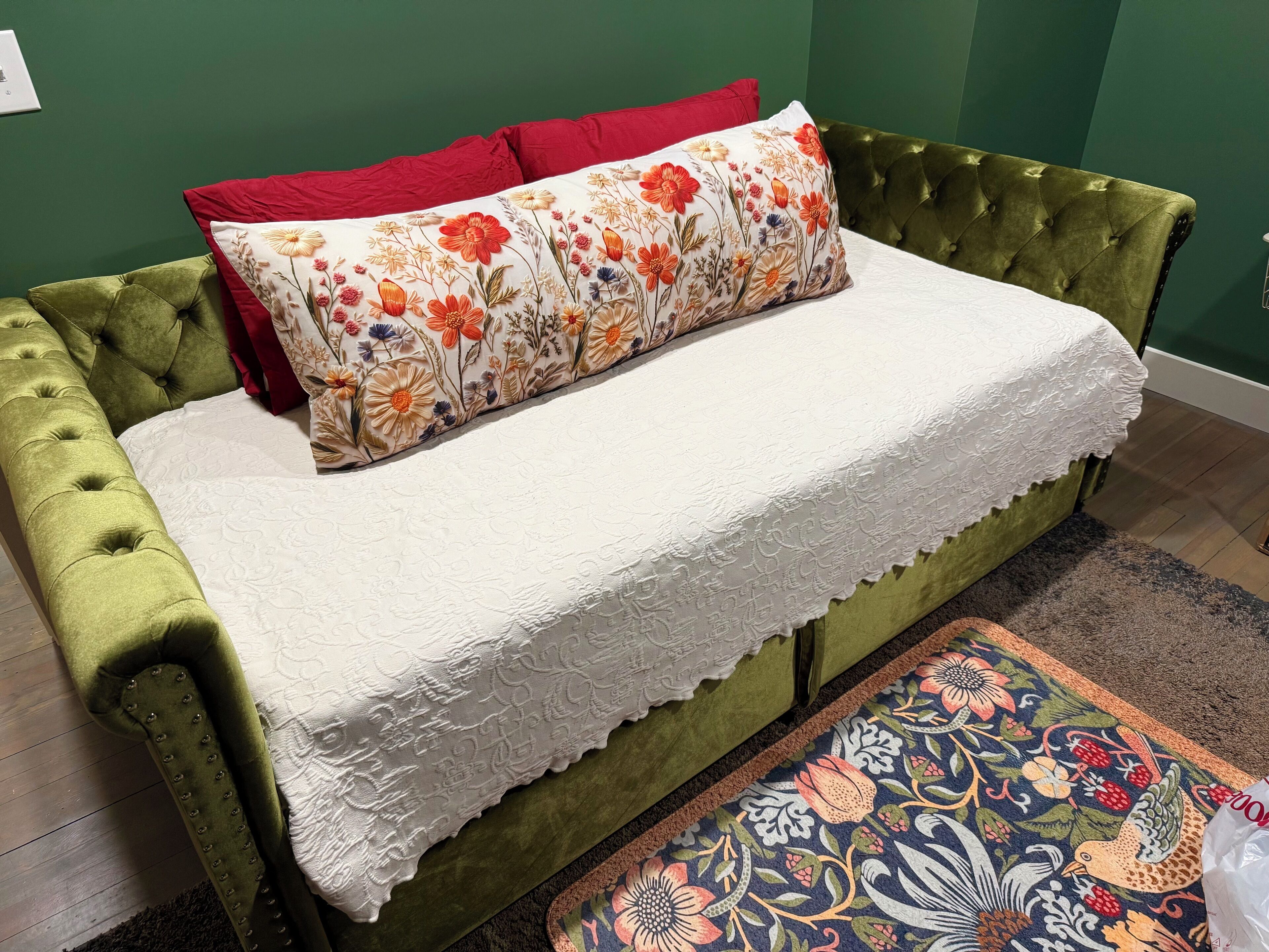 Un sofá cama verde con tufos, una colcha de encaje blanco y una almohada estampada.