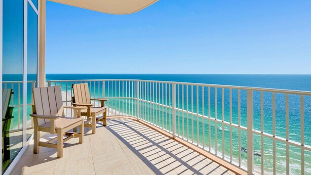 A balcony with two wooden chairs and a table, overlooking a clear blue sea. - NEW Phoenix Gulf Tower II Unit 2508 | Luxury East Corner - Gorgeous Coastal Decor, Stunning Views (Orange Beach)