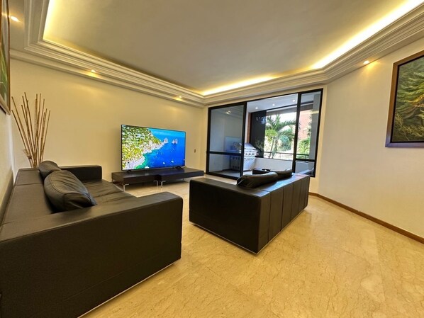 Ein modernes Wohnzimmer mit einem Flachbildfernseher, schwarzen Sofas und einem großen Fenster mit Blick ins Freie.