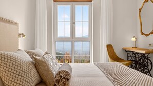 Ein Schlafzimmer mit großem Fenster, einem Bett mit gemusterten Textilien, einem Sessel, einem Tisch und einem Spiegel.
