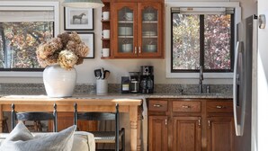 A modern kitchen with wooden cabinets, a stainless steel refrigerator, a dining area with a bench and cushions, and a large window with a view of trees. - Par & Pine Lodge | Double decks w/ fire pit! (Basye)