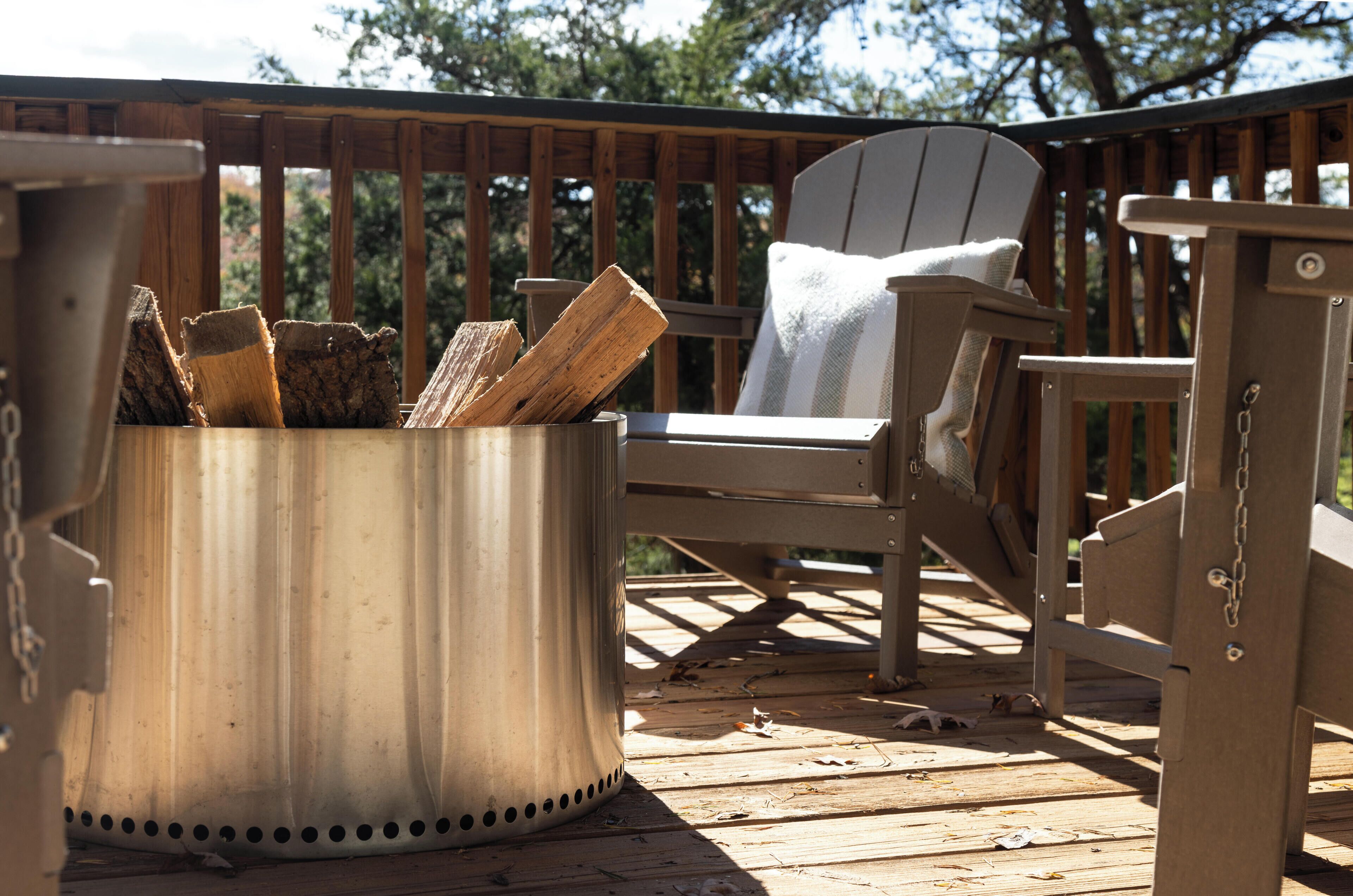 A wooden deck with a metal fire pit containing logs, a white chair, and a wooden table.