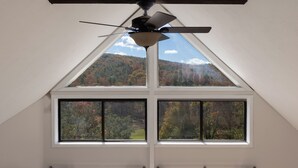 A room with a sloped ceiling, a ceiling fan, and two large windows offering a view of a forested landscape. - Par & Pine Lodge | Double decks w/ fire pit! (Basye)