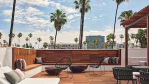 Un salón en la azotea con suelo de madera, asientos acolchados y vistas a palmeras y edificios.