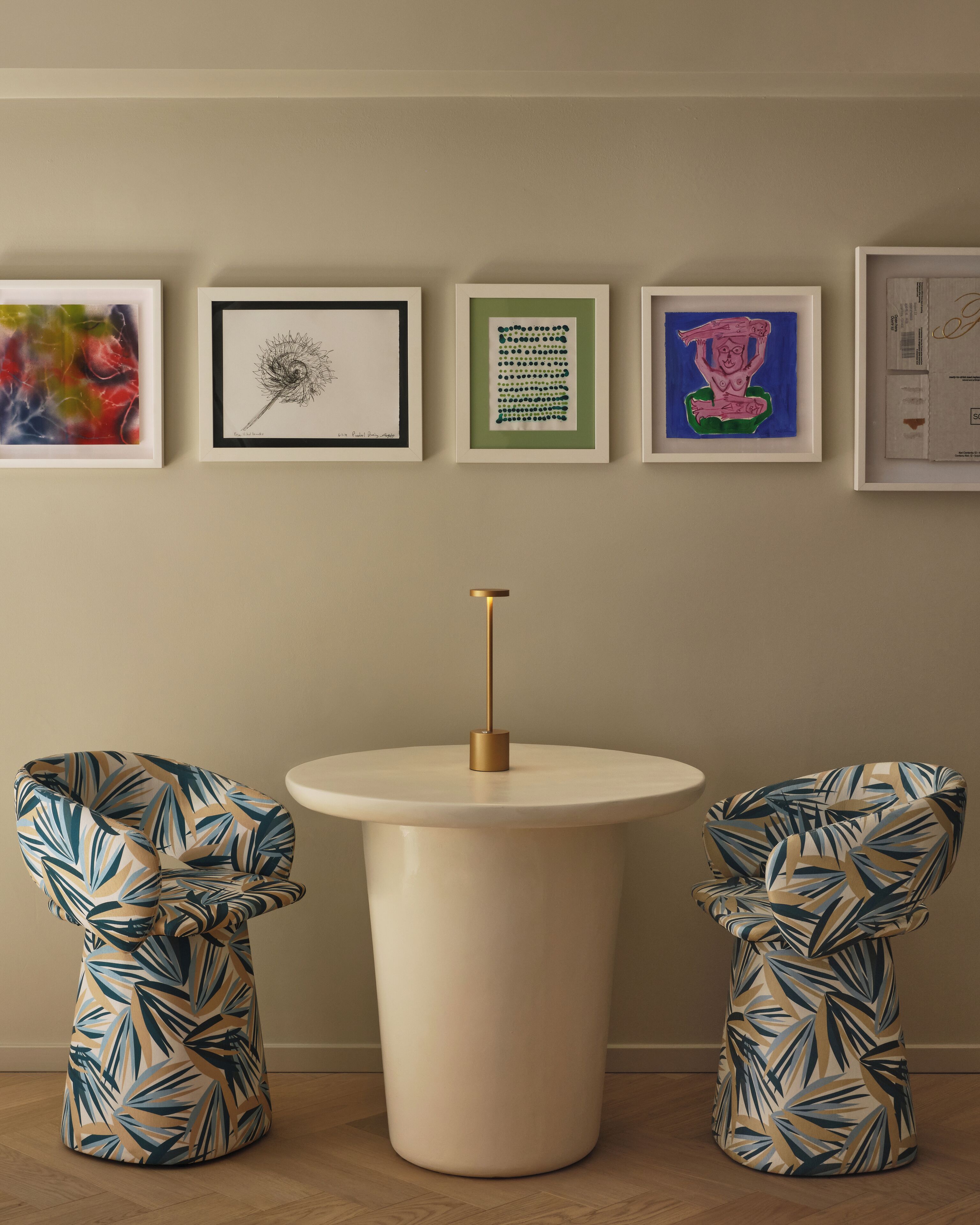 Une pièce comprenant une table ronde, deux chaises ornées d’un motif de feuilles tropicales et quatre œuvres d’art encadrées accrochées au mur.