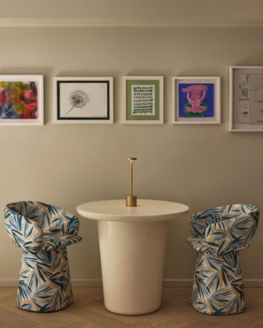 A room with a round table, two chairs with a tropical leaf pattern, and four framed artworks on the wall. - The Ivory (Los Angeles)