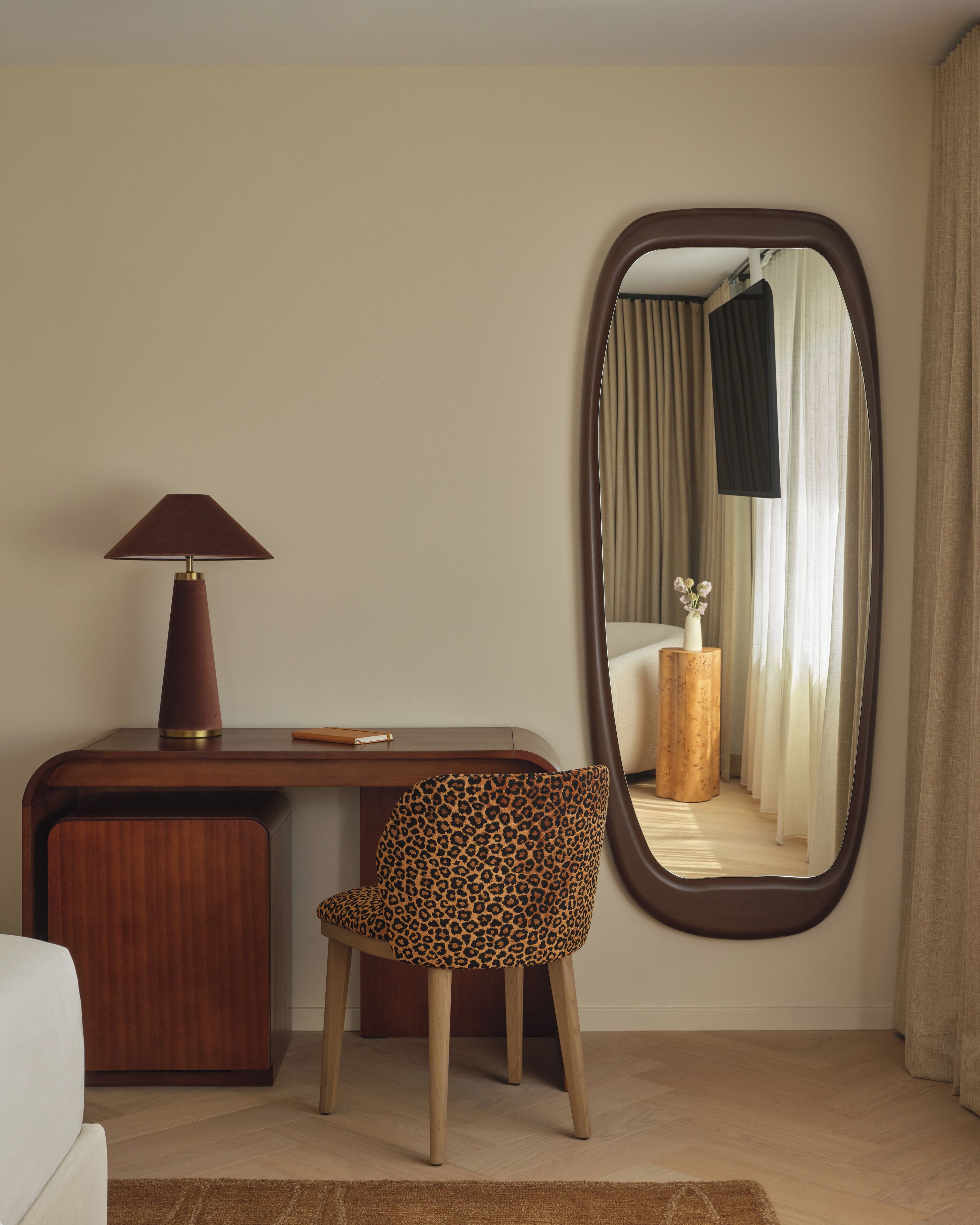 A room with a desk, a chair, a lamp, and a mirror reflecting a bed and curtains.