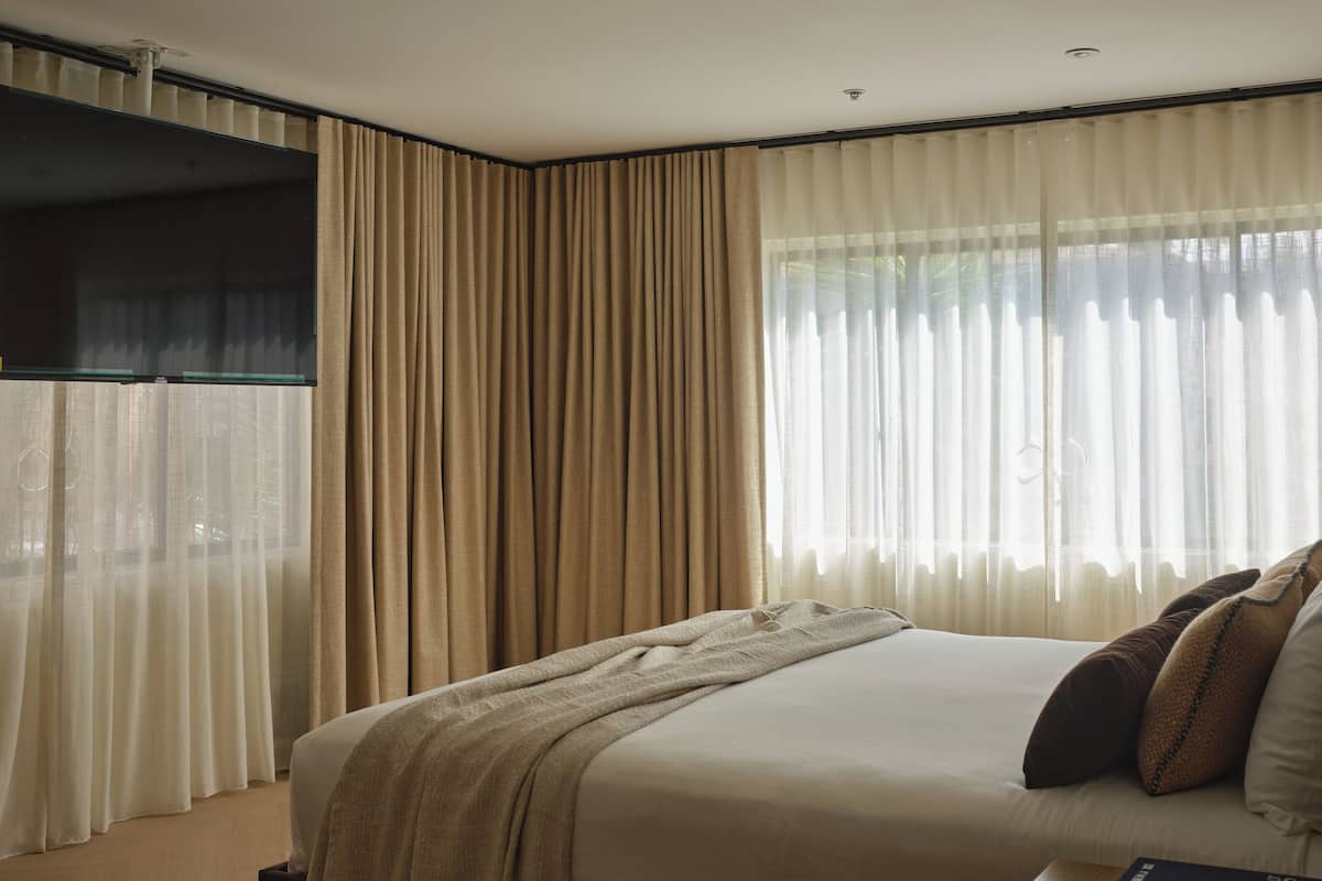 Une chambre d’hôtel avec un grand lit, des rideaux beiges, une fenêtre avec des rideaux en voile et un téléviseur à écran plat fixé au mur.