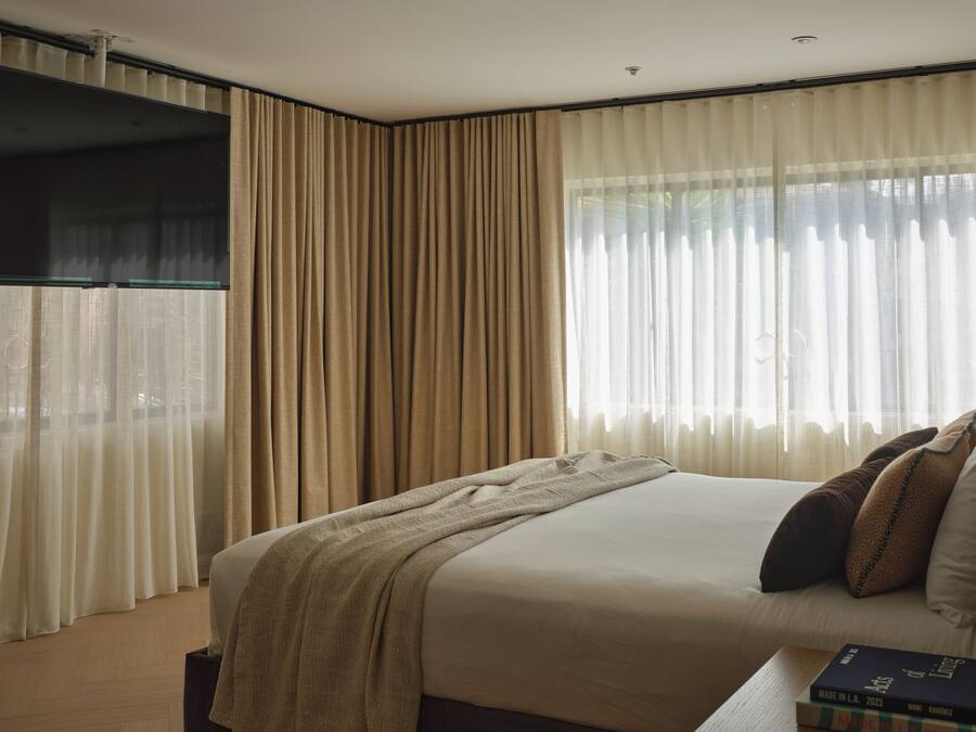 Une chambre d’hôtel avec un grand lit, des rideaux beiges, une fenêtre avec des rideaux en voile et un téléviseur à écran plat fixé au mur.