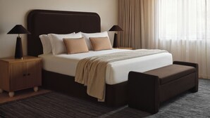 Habitación de hotel con cama, dos mesitas de noche, un banco, cabecera y ventana con cortinas.