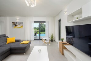 Ein modernes Wohnzimmer mit grauem Sofa, einem weißen Couchtisch, einem Flachbildfernseher und Blick auf einen Swimmingpool durch die Glastür.