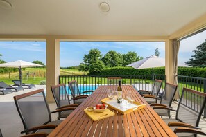Ett träbord med sex stolar, en vinflaska och en skål med frukt står på en terrass med utsikt över en swimmingpool och grönområden.