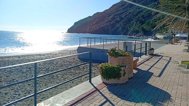 En kystpromenade med gelænder, plantekasser og udsigt til havet og en bakke.