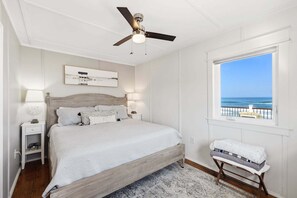 A bedroom with a bed, a ceiling fan, a nightstand, a bedside table, a window with a view of the ocean, and a bench by the window. - Night Heron: Oceanfront Views + Walk to Dining (Kitty Hawk)