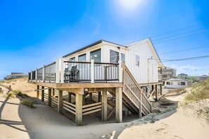 Una casa sulla spiaggia con una veranda in legno e un balcone, sollevata su palafitte sopra la sabbia.