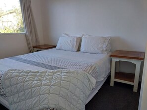 Ein ordentlich bezogenes Bett mit einem gemusterten Bettüberwurf und Kissen, ein Nachttisch und ein Fenster mit Vorhängen.