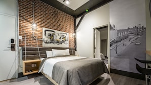 A modern bedroom with a brick wall, a bed with a grey bedspread, a hanging light fixture, and a large black and white cityscape mural on the wall. - La Suite Hotel (Shawinigan)