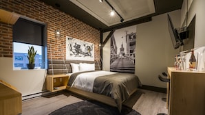 A modern bedroom with a brick wall, a large bed, a desk, and a window with a plant. - La Suite Hotel (Shawinigan)
