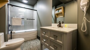 A bathroom with a toilet, shower, vanity with a mirror, and a hairdryer mounted on the wall. - La Suite Hotel (Shawinigan)