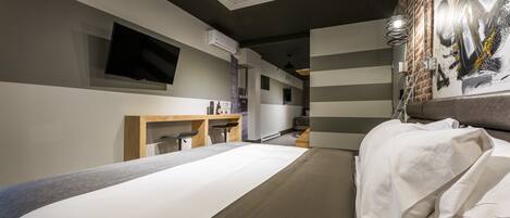 A modern hotel room with a large bed, a flat-screen TV mounted on the wall, a desk with a chair, and a brick wall with a painting.