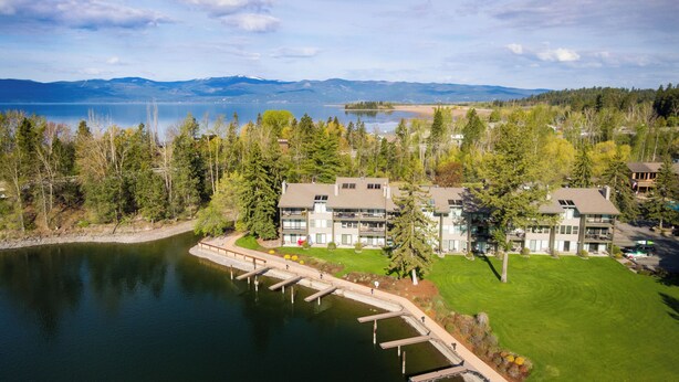 A lakeside hotel with a dock, a well-maintained lawn, and a view of mountains in the distance. - New! Patio, AC & Classic Comfort! (Bigfork)