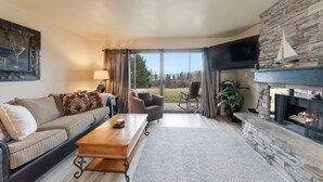A living room with a stone fireplace, a sofa, a coffee table, and a television. - New! Patio, AC & Classic Comfort! (Bigfork)