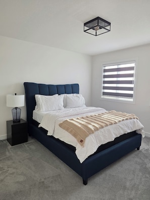 Een slaapkamer met een donkerblauwe bed, witte beddengoed, een bedlamp en een raam met zonwering.
