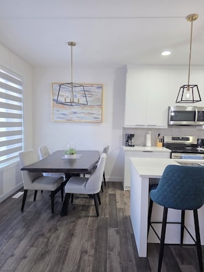 Een moderne keuken met witte kasten, een inbouwmagnetron en een eethoek met een donker houten tafel en grijs gestoffeerde stoelen.