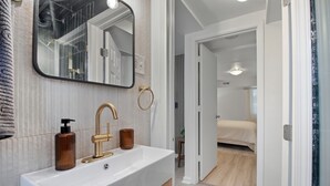 Une salle de bain avec un lavabo blanc, un robinet doré et un miroir. Il y a un porte-serviette au mur et un flacon de savon.