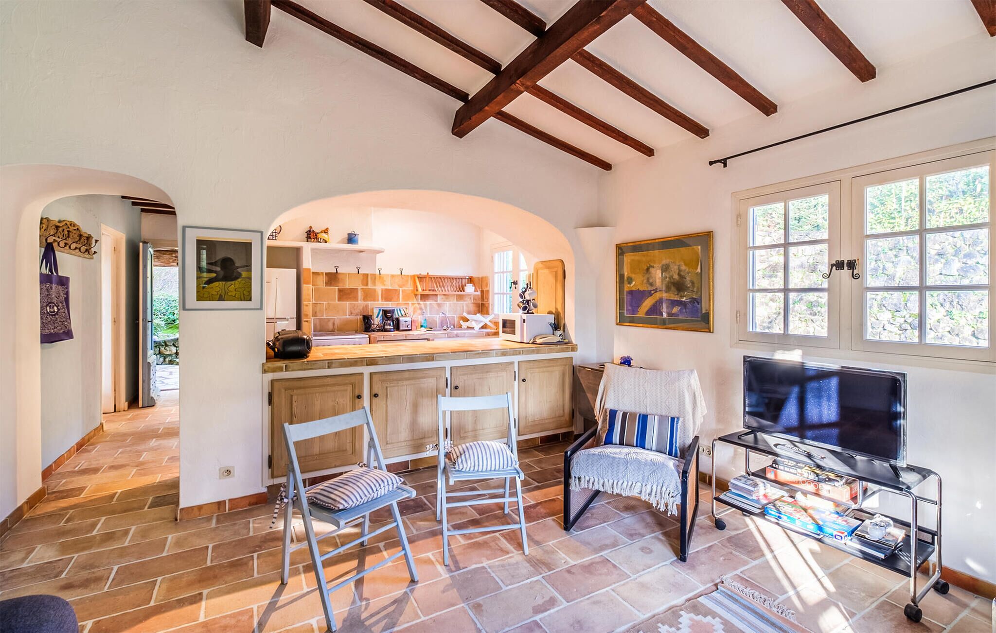 A spacious kitchen with wooden beams, a dining area with chairs, a TV on a cart, and a large window.