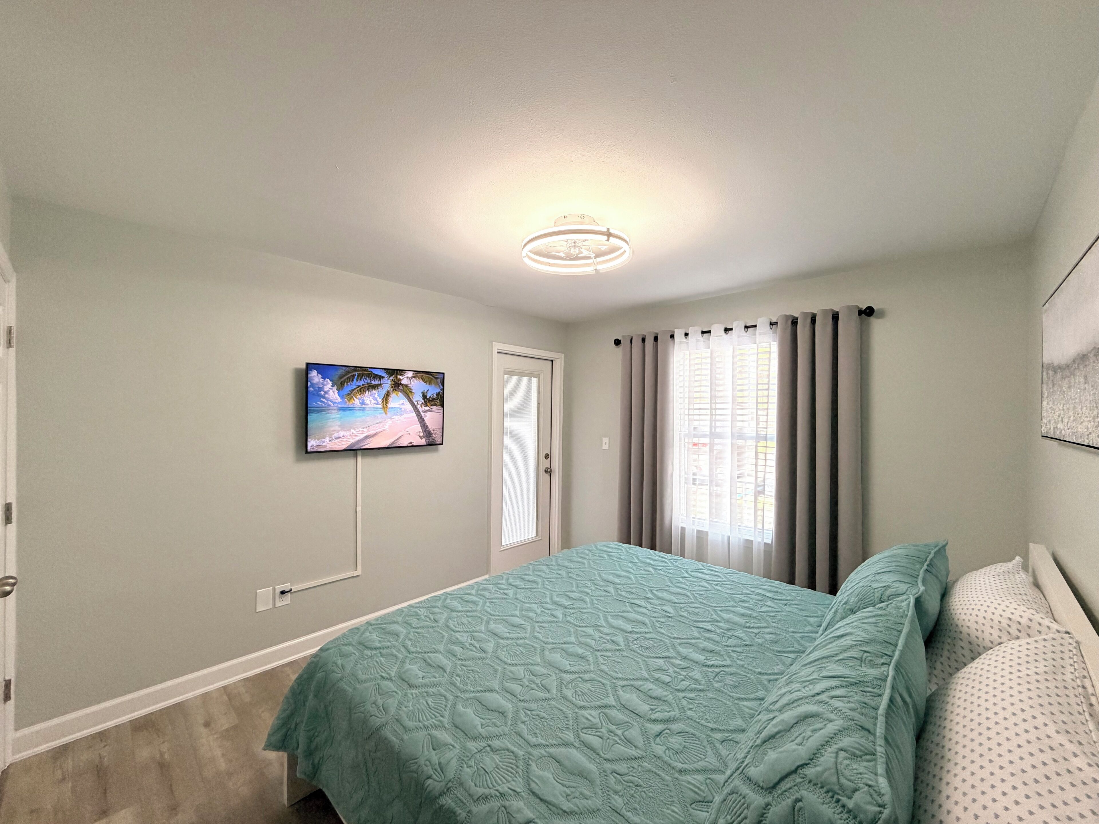 Un dormitorio con cama, televisor en la pared, ventana con cortinas y puerta.