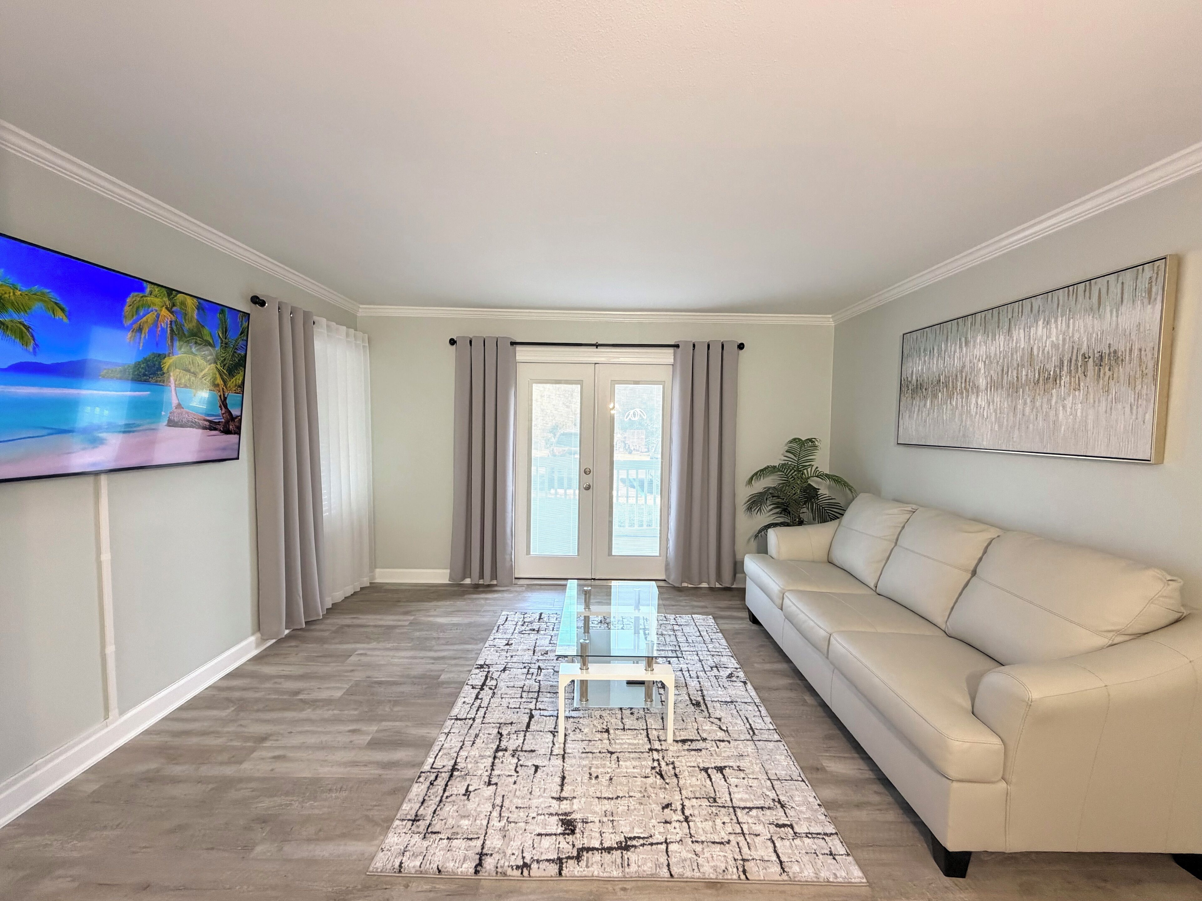 A living room with a beige sofa, a glass coffee table, a patterned rug, a flat-screen TV, and a large abstract painting on the wall.