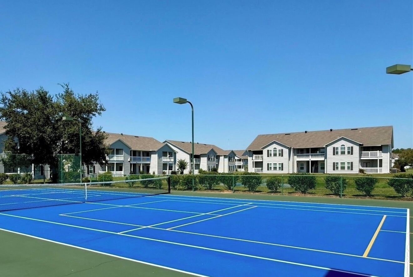 A tennis court with a net in the middle, surrounded by a fence. There are buildings with multiple stories in the background.