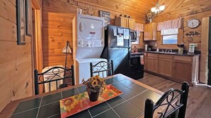 A cozy kitchen with wooden walls, a dining table with chairs, a refrigerator, a microwave, and a clock on the wall. - Moonlight Ridge Cabin (Bryson City)