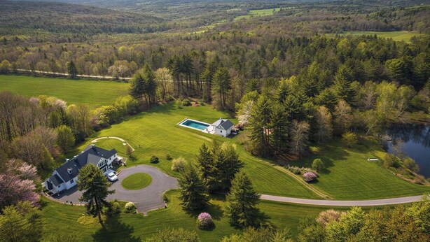 Vue aérienne d’une propriété comprenant une grande maison, une piscine et un lac entouré d’une forêt dense.