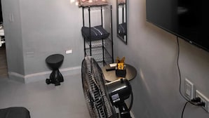 Ein kompakter Wohnbereich mit Wandfernseher, Standventilator, Hocker und einer Regalwand mit Spiegel.